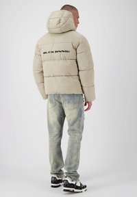 Beige gewatteerde jas met een capuchon, voorzien van een logo op de rug, gecombineerd met lichte spijkerbroek en zwarte sneakers in een minimalistische setting.