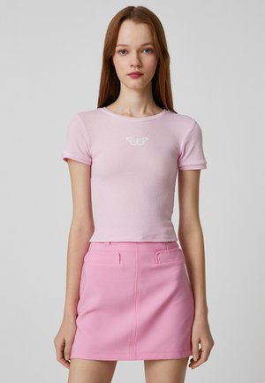 Roze ribgebreide top met korte mouwen en een vlinderontwerp op de borst, gecombineerd met een roze minirok met twee voorzakken en strakke lijnen.