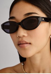 Óculos de sol pretos em formato de olho de gato com lentes escuras, armação grossa em plástico brilhante e detalhes em dourado subtil. Design minimalista, textura suave.