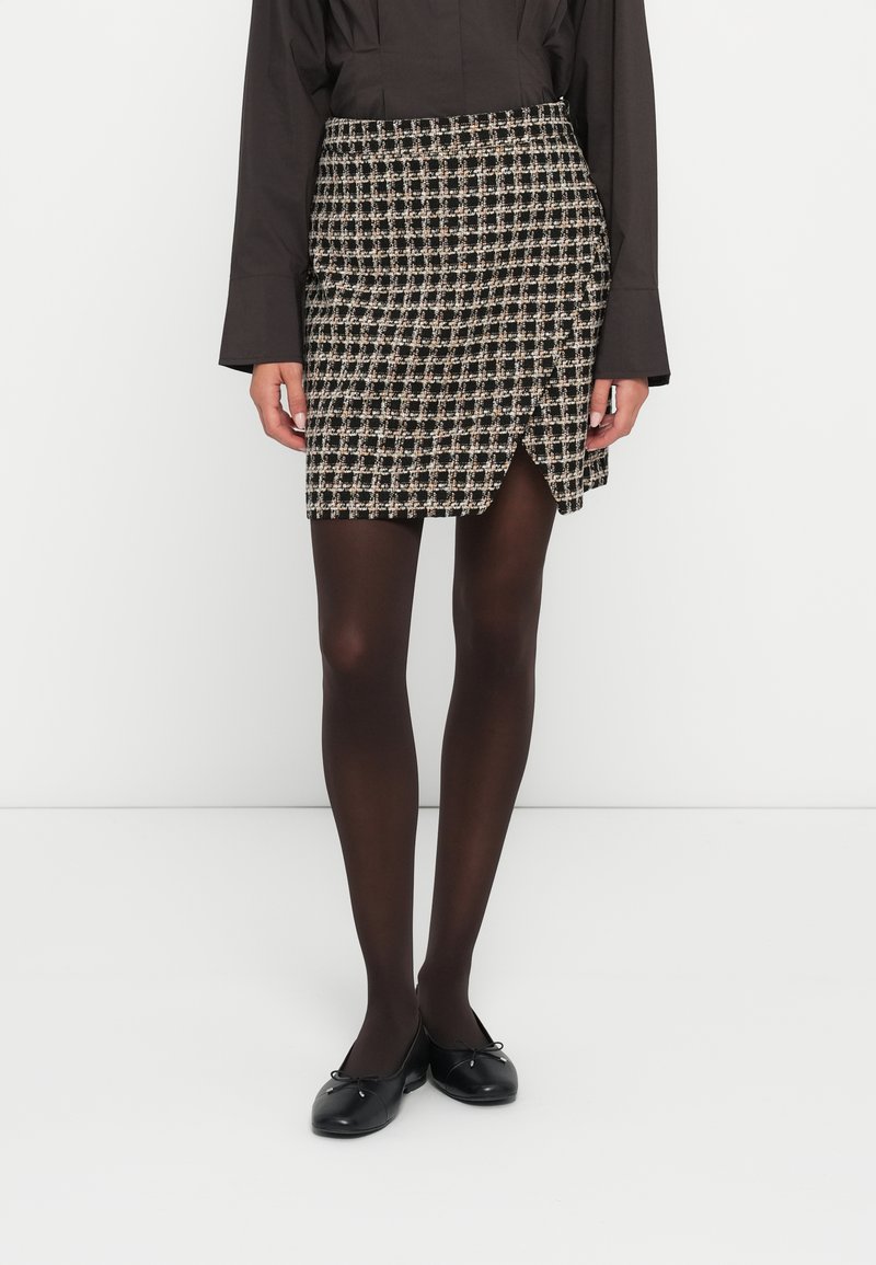 Falda hasta la rodilla con un patrón de cuadros en negro y beige, que presenta un diseño de envoltura frontal, hecha de tela texturizada, combinada con medias negras.