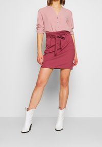 Žena v růžové blůze, s vysokým pasem vínově červené sukně se stahovací šňůrkou a bílými botami s špičatou špičkou do kotníků, stojí na neutrálním pozadí.