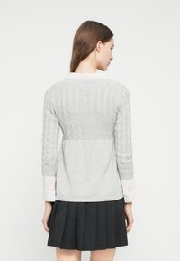 Γυναίκα με μακριά καστανά μαλλιά που φοράει ένα ανοιχτό γκρι πουλόβερ τύπου καλώδιο πάνω από μια ριγέ μπλούζα και μια σκούρα πλισέ φούστα, γυρισμένη προς τα πίσω.