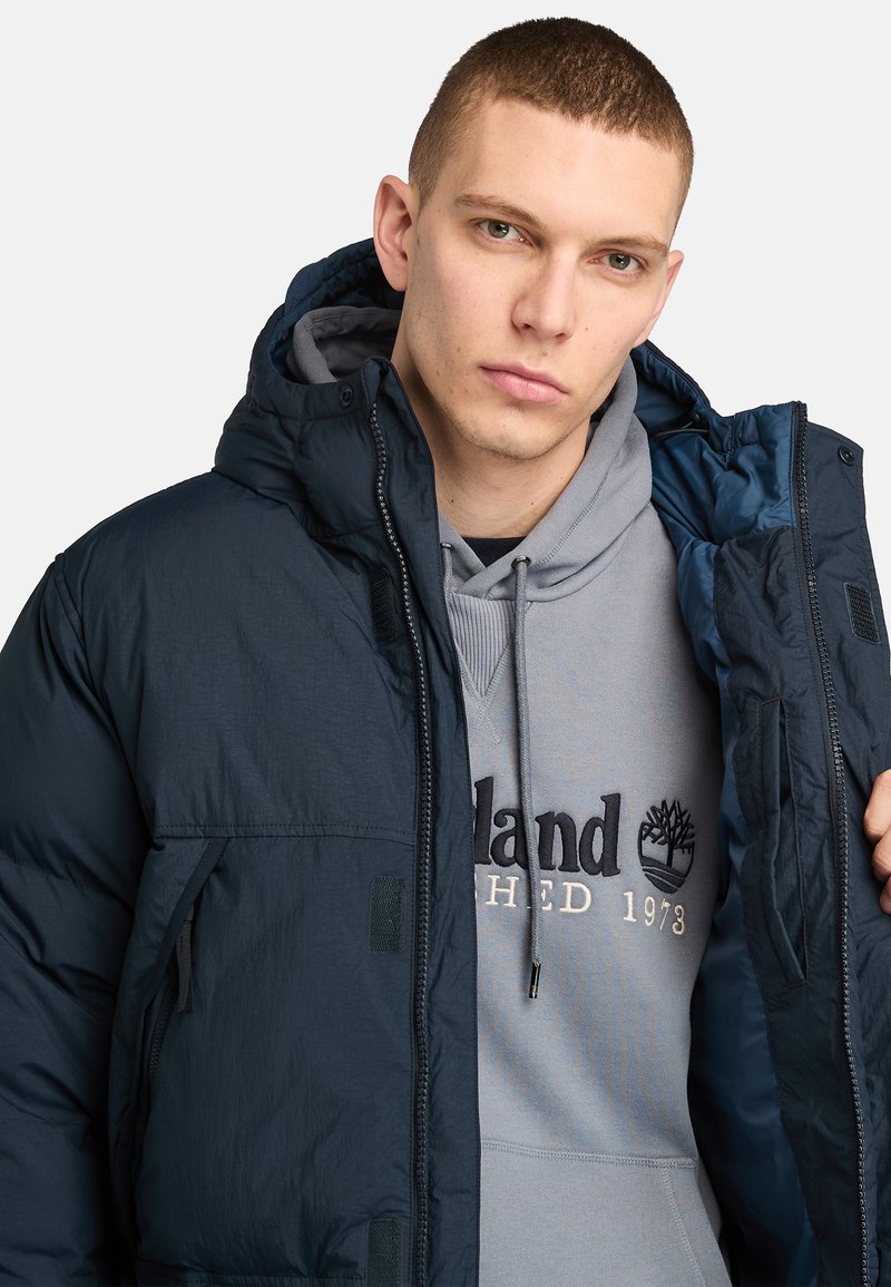 Marineblaue Pufferjacke mit strukturierter Oberfläche, ausgestattet mit einer Kapuze und Reißverschlusstaschen. Getragen über einem grauen Hoodie mit Logo.