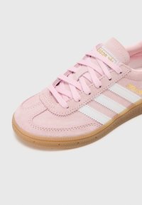 Sapatilhas de camurça rosa claro com riscas brancas, bico redondo, sola de borracha e atacadores rosa claro. Apresenta uma marcação subtil em dourado na lateral.