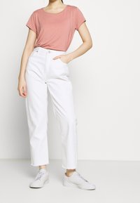 Lys rosa t-shirt med korte ærmer parret med højtaljede hvide vidde jeans og hvide sneakers. Blødt stof og slidt kant på jeansene.