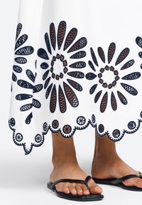 White dress with navy embroidery featuring floral patterns and scalloped hem. Black thong sandals on bare feet below the dress.