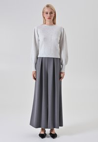 Donna in piedi contro uno sfondo neutro, che indossa un maglione bianco a maniche a sbuffo, pantaloni grigi larghi e plissettati, e scarpe nere con punte a forma di triangolo decorate con fiocchi.