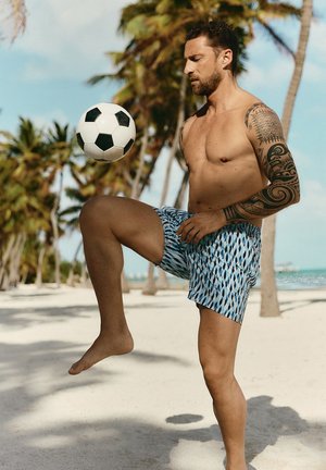 Mężczyzna bez koszulki z tatuażami żonglujący piłką nożną na piaszczystej plaży z palmami i niebieskim niebem w tle.