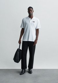 Mann i hvit polo og svarte bukser, holder en svart veske i venstre hånd, står mot en ensfarget grå vegg og gulv.