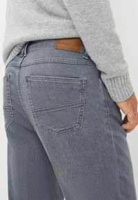 Uomo che indossa un maglione grigio a maglia e jeans blu sbiaditi con un'etichetta in pelle marrone sulla cintura posteriore, mano appoggiata di lato.