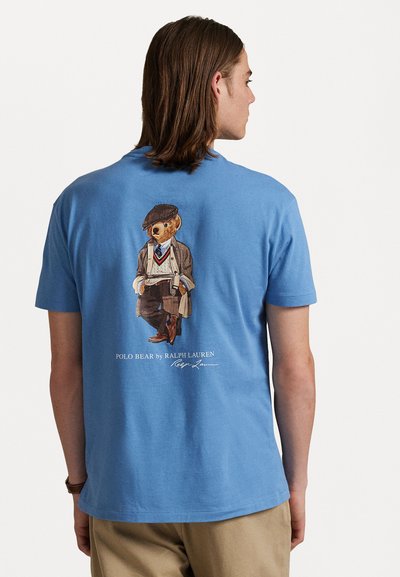 Tricou bleu deschis din bumbac, având un grafic cu un urs îmbrăcat în costum pe spate, cu textul "Polo Bear by Ralph Lauren" dedesubt.