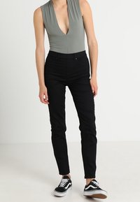 Grå ærmeløs bodysuit med dyb V-udskæring, parret med sorte slim-fit bukser og sorte sneakers med hvide såler. Glat stofstruktur.