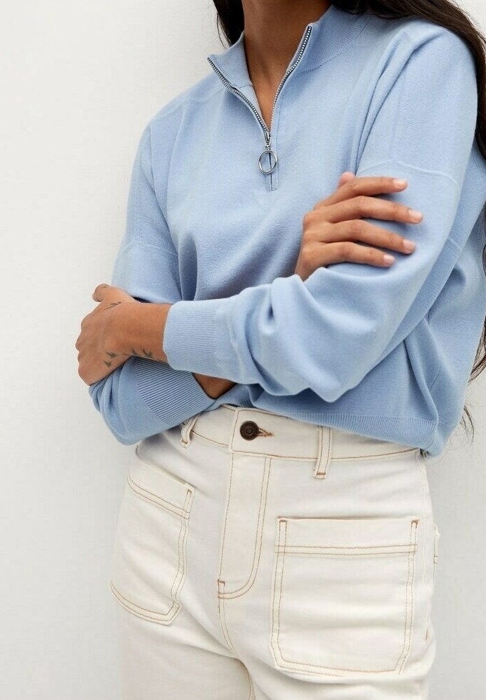 Suéter azul claro con cremallera y cuello alto, con herrajes plateados, combinado con jeans blancos de cintura alta con costuras en contraste.