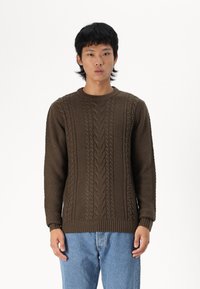 Suéter marrón de punto con patrones de trenzado y textura, con cuello redondo y puños acanalados. Combinado con jeans de mezclilla azul claro.