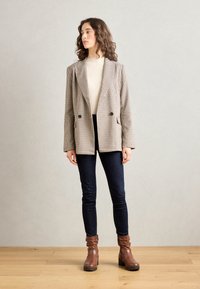 Blazer en un patrón de jaquard marrón y crema, con un frente de doble botonadura y solapas con muescas, combinado con jeans oscuros ajustados y botas marrones.