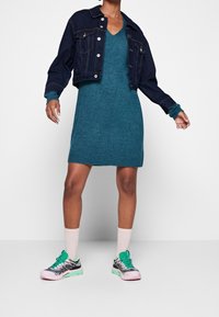 Giacca cropped in denim scuro sopra un vestito in maglia teal. Abbinata a sneakers pastello e calzini rosa chiaro. Mostra uno stile rilassato e stratificato.
