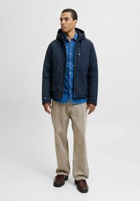 Veste imperméable marine avec fermeture éclair et boutons, portée sur une chemise en denim. Un pantalon large beige et des chaussures sombres complètent la tenue.