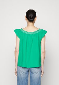 Top verde, solto, com mangas a bailar e decote bordado. O tecido tem uma textura suave; a vista de trás mostra uma silhueta descontraída.