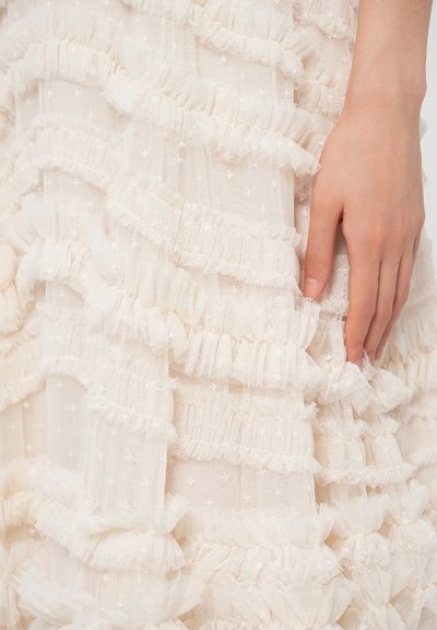 Το λευκό ύφασμα με βολάν αποτελείται από στρώσεις διάφανου υλικού με μικρά αστέρια. Ένα χέρι αγγίζει την υφή, υπογραμμίζοντας τη λεπτομέρεια.