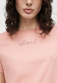 Γυναίκα με καστανά σγουρά μαλλιά που φοράει ανοιχτό ροζ πουκάμισο με μικρές λεπτομέρειες από κοσμήματα που σχηματίζουν κυκλικό κείμενο στο στήθος.