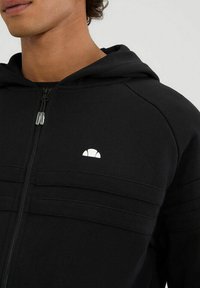 Sudadera negra con cremallera hecha de un tejido suave, que presenta un distintivo logo blanco en el pecho y franjas texturizadas horizontales en el cuerpo.