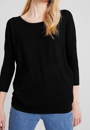 Frau trägt einen schlichten schwarzen Langarmpullover mit Rippstrickärmeln und blaue Jeans vor neutralem Hintergrund.