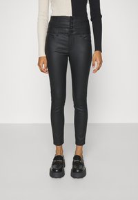 Kvinna klädd i höga svarta slim-fit byxor med fyra knappar, svarta kraftiga loafers och en långärmad tröja med färgblocker.