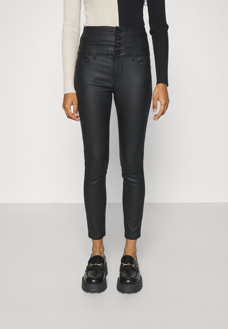 Kvinna klädd i höga svarta slim-fit byxor med fyra knappar, svarta kraftiga loafers och en långärmad tröja med färgblocker.