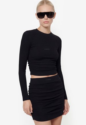 Žena v čiernom dlhom rukáve s krátkym topom a riasenou minisukňou so slnečnými okuliarmi, stojaca pred jednoduchým bielym pozadím.