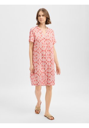Vrouw loopt, draagt een knielange, koraal wit met patroon jurk met korte mouwen en beige sandalen tegen een effen lichte achtergrond.