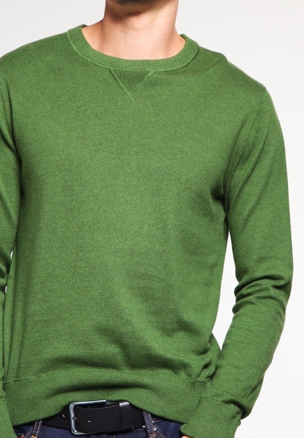 Homme portant un pull vert uni à manches longues rentré dans un jean bleu avec une ceinture noire sur un fond blanc.
