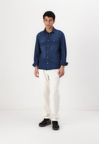 Camisa de mezclilla en azul oscuro con mangas largas, cierre de botones en la parte delantera y dos bolsillos en el pecho. Lleva puesta con pantalones blancos y zapatillas negras.