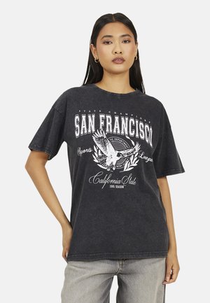 Woman wearing a black oversized T-shirt with "San Francisco" text and eagle graphic, paired with light gray jeans against a plain background.
