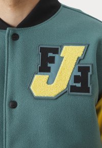 Green fleece varsity jacket featuring a black ribbed collar, bold yellow and black appliqué letters "J" and "F," and textured accents.