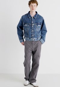 Denim jakna s klasičnim krojem v modri barvi, z sprednjimi žepi in srebrnimi gumbi, v kombinaciji s sivo cargo hlačami z stranskimi žepi.