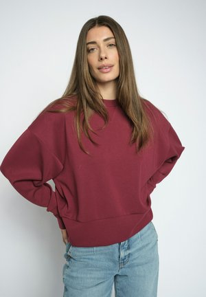 Femme aux longs cheveux bruns portant un sweat-shirt ample bordeaux et un jean bleu clair sur un fond clair uni.