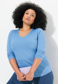 Světle modrý top s dlouhým rukávem a výstřihem do V, s nařasenými rukávy po tři čtvrtiny, v kombinaci s prošívanou levandulovou kabelkou, na bílém pozadí.