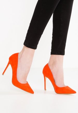 Oranssi mokkanahasta valmistetut korkokengät, joissa on teräväkärkinen muotoilu ja stilettokorko. Yhdistetty mustiin kapeisiin housuihin, mikä korostaa kengän eläväistä väriä ja muotoilua.