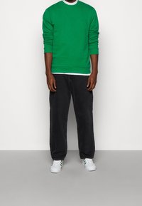Groene crewneck sweatshirt met lange mouwen, gedragen over een witte top, gecombineerd met zwarte relaxed-fit jeans en witte sneakers met groene accenten.