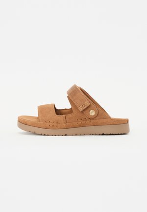 Brune semskede sandaler med åpen tå, justerbar stropp, teksturert såle og en sirkulær logoakseent på siden.