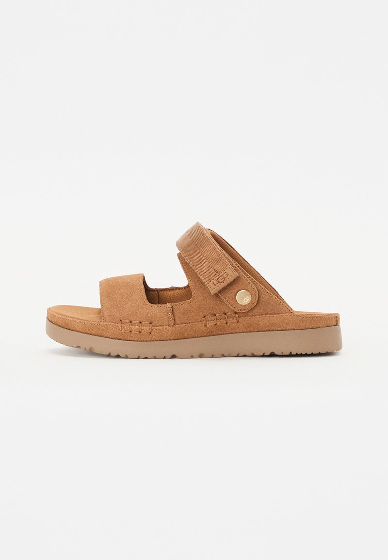 Pruunist nubukist libistav sandaal, millel on reguleeritav rihm, avatud varvaste disain, tekstureeritud põhi ja ringikujuline logoaktsent küljel.