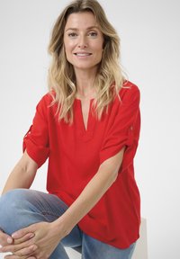 Blusa rossa con scollatura a V e maniche arrotolate, realizzata in tessuto leggero. Indossata con jeans blu, caratterizzati da cuciture visibili e texture liscia.