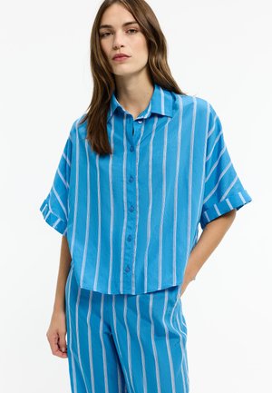 Woman wearing a loose blue button-up shirt with white vertical stripes and matching striped pants, standing with one hand in pocket.