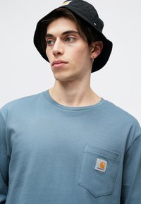 Niebieska koszula z długim rękawem z kieszonką z przodu, z logo Carhartt. Noszona z czarną czapką typu bucket hat. Gładka tekstura materiału.