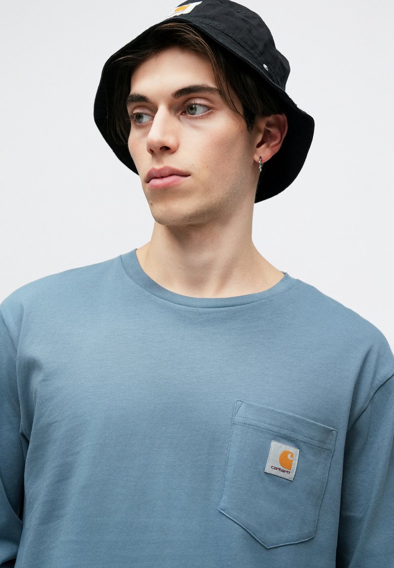 Blå langærmet skjorte med en frontlomme, prydet med et Carhartt-logo. Båret med en sort bøjlestråhat. Glat stoftekstur.