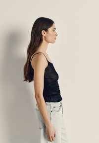 Jauna sieviete melnā bezroku topā un gaiši zilos džinsos stāv profilā pret vienkāršu bēša krāsas fonu.