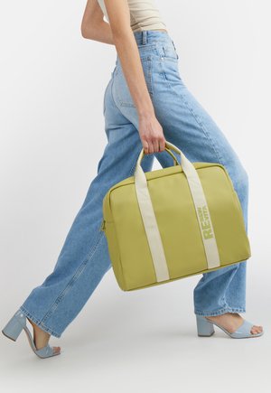 Donna con jeans blu e tacchi azzurri chiaro, che porta una grande borsa tote gialla con manici crema e scritta "NEW RELM".
