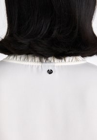 Balta blūze ar viļņotu apkakli un melnu dekoratīvu pogu aizmugurē. Gluda tekstūra ar vienkāršu un mūsdienīgu dizainu.