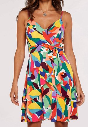Femme portant une robe sans manches à col en V avec un motif abstrait de feuilles multicolores et une taille nouée, sur un fond uni.