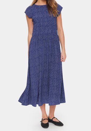 Femme portant une robe midi bleue à taches blanches et manches courtes, associée à des chaussures plates noires avec des brides à la cheville.